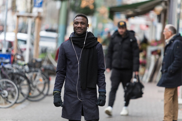 Young Black man listening to earphones standing on sidewalk with incidental people. Focus on foreground. - Фото, изображение