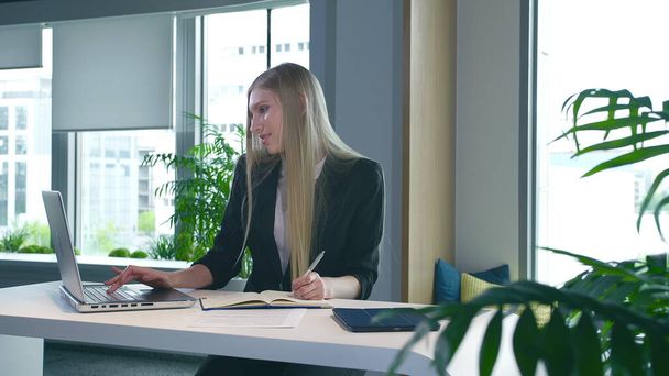 Elegant woman working in stylish office. Modern blond woman in trendy suit sitting at table in light contemporary office and writing on papers. - Photo, Image