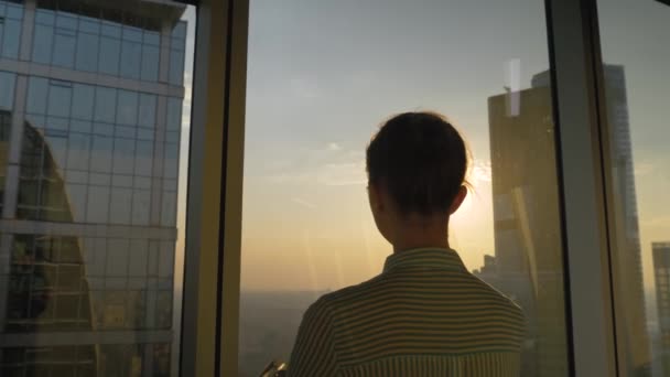 Rückansicht einer Frau, die durch das Fenster eines Wolkenkratzers den Sonnenuntergang betrachtet - Filmmaterial, Video