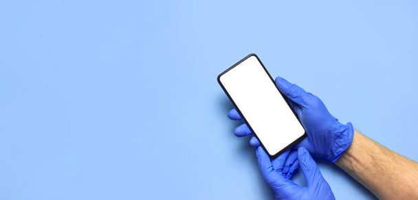 Male hands in blue medical gloves hold smartphone with white blank screen on blue background. Protecting Health During a Pandemic coronavirus 2019-nCoV Hands medical worker volunteer doctor with phone. - Photo, Image