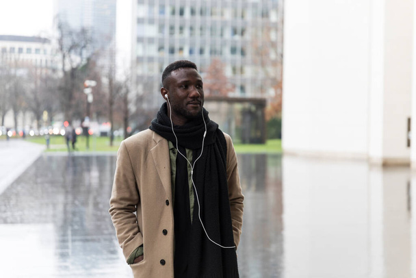 Young black man in beige coat and listening to earphones walking in city and looking away.  - Photo, image