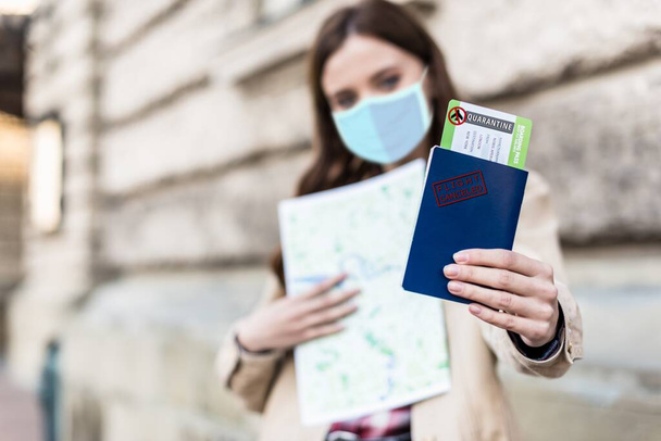 Selective focus of woman in medical mask holding map and showing passport and air ticket with quarantine illustration - Photo, Image