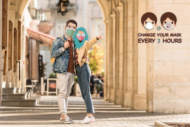 Tıbbi maskeli, sokakta kucaklaşan ve parmağıyla işaret eden iki turist her 3 saatte bir maskenizi değiştirin. - Fotoğraf, Görsel