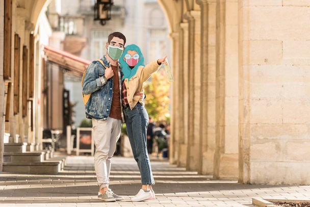 Paar Touristen mit abgebildeten Gesichtern in medizinischen Masken umarmen sich auf der Straße und zeigen mit dem Finger weg - Foto, Bild