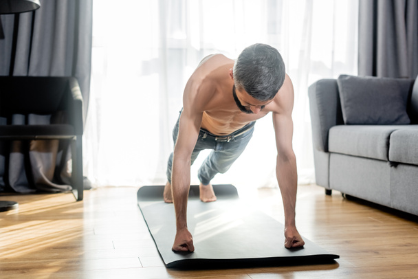 Shirtless man doing press ups while training on fitness mat in living room - Zdjęcie, obraz