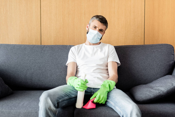 Man in medical mask holding rag and detergent while sitting on couch  - Foto, immagini