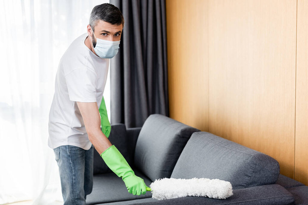 Άντρας με ιατρική μάσκα και γάντια καθαρισμού καναπέ με βούρτσα σκόνης  - Φωτογραφία, εικόνα