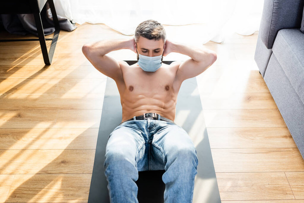 Hochwinkelaufnahme eines muskulösen Mannes in medizinischer Maske beim Bauchmuskeltraining auf der Fitnessmatte im Wohnzimmer - Foto, Bild