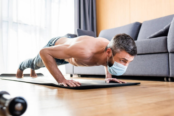 Selective focus of shirtless man in medical mask doing press ups on fitness mat in living room - Foto, Imagem