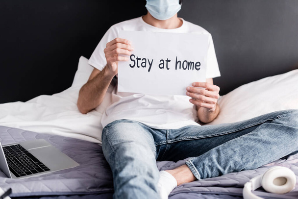 Vista ritagliata dell'uomo in maschera medica con carta con soggiorno a casa lettering vicino laptop e cuffie sul letto
  - Foto, immagini