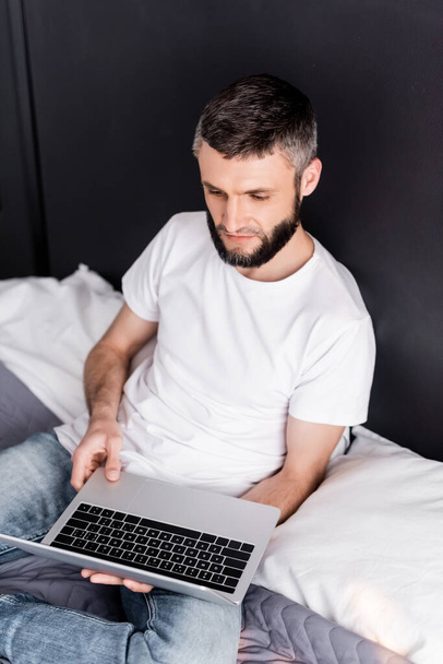 Handsome freelancer holding laptop while sitting on bed  - Photo, Image