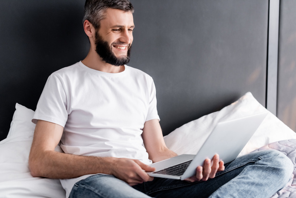 Handsome freelancer smiling while holding laptop on bed  - Фото, изображение