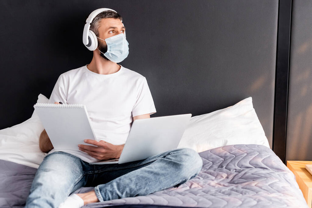 Фрилансер в медицинской маске и наушниках, работающий с ноутбуком и ноутбуком на кровати
  - Фото, изображение
