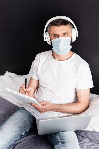 Freelancer in headphones and medical mask writing on notebook near laptop on bed  - Фото, изображение