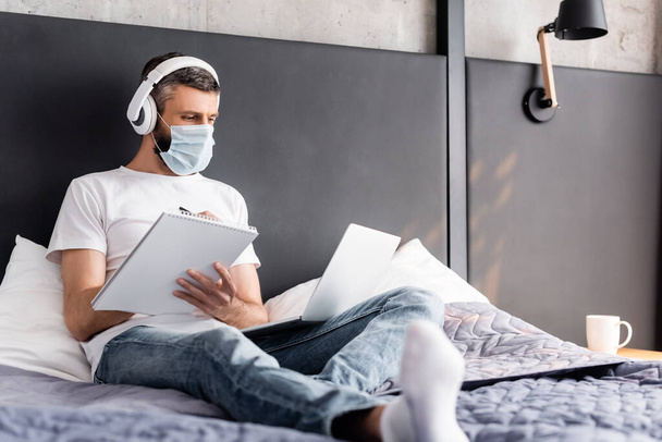 Selective focus of teleworker in headphones and medical mask whiting on notebook and using laptop in bedroom - Photo, Image