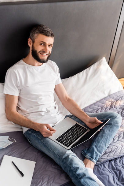 High angle view of smiling man holding credit card and laptop near medical mask and notebook on bed  - Fotografie, Obrázek