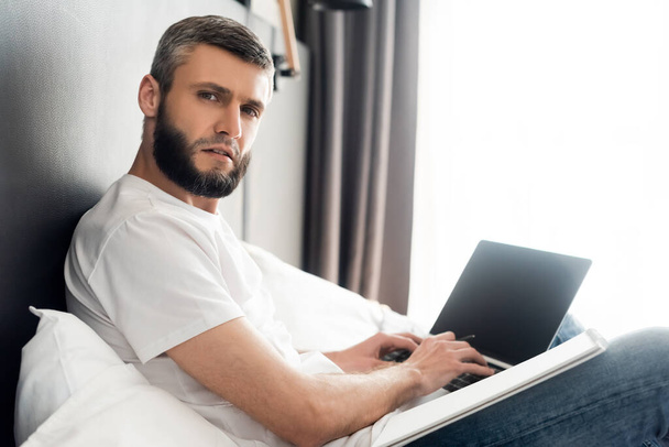 Side view of handsome freelancer looking at camera while using laptop on bed  - Photo, Image