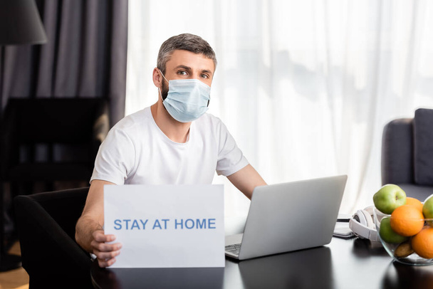 Freelancer in medisch masker holding card met verblijf thuis belettering in de buurt van laptop en fruit op tafel  - Foto, afbeelding