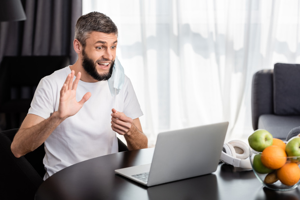 Smiling freelancer having video call on laptop and holding medical mask at home  - Foto, Bild