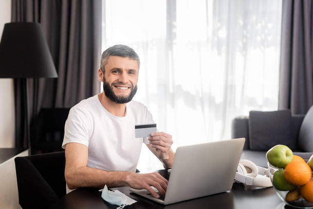 Усміхнений телефонний працівник тримає кредитну картку і використовує ноутбук біля медичної маски та фруктів на столі
  - Фото, зображення