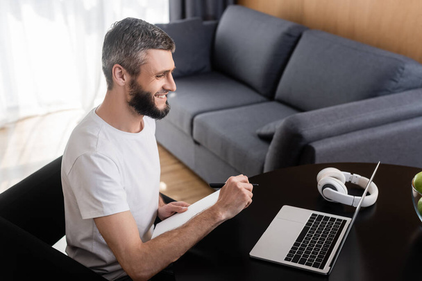 Side view of smiling freelancer holding notebook near laptop and headphones on table at home  - Photo, Image