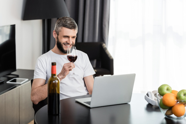 Selective focus of smiling freelancer holding glass of wine near laptop and fruits on table  - Zdjęcie, obraz