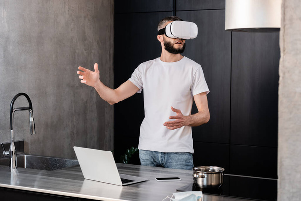 Selective focus of man using vr headset near gadgets and medical mask on kitchen worktop  - Foto, afbeelding