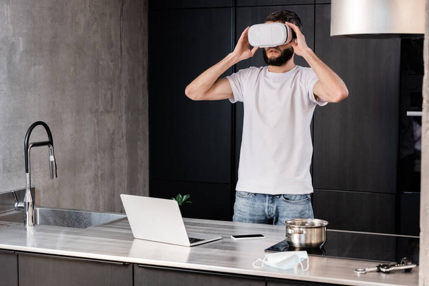 Man met behulp van virtual reality headset in de buurt van digitale apparaten en medisch masker op keukenblad  - Foto, afbeelding