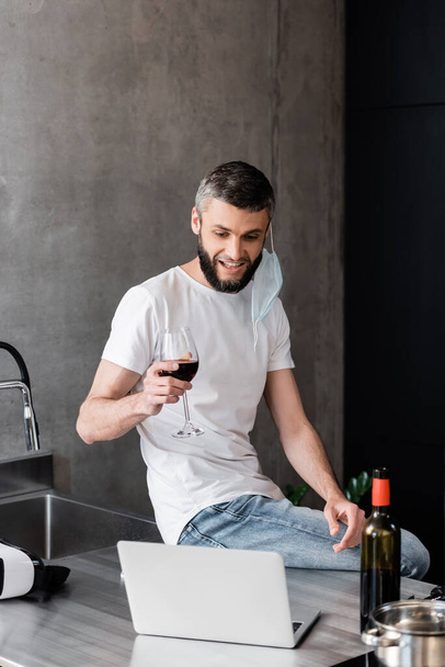 Smiling man in medical mask holding glass of wine near laptop and vr headset on worktop in kitchen  - Photo, Image