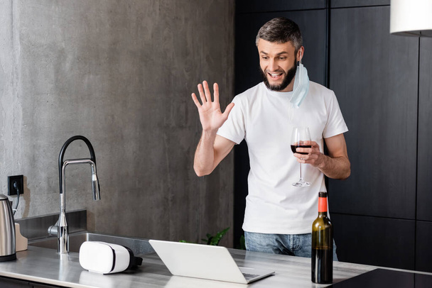 Smiling man in medical mask holding glass of wine while having video call on laptop in kitchen  - Foto, imagen