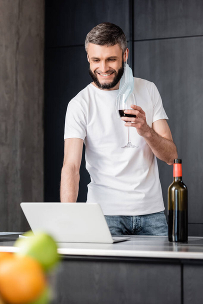 Selective focus of smiling man in medical mask holding glass of wine and using laptop in kitchen  - Foto, immagini