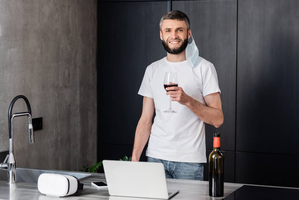 Man in medical mask holding glass of wine and smiling at camera near laptop on worktop in kitchen - Valokuva, kuva