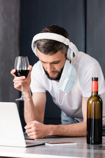 Selective focus of freelancer in medical mask and headphones holding glass of wine near gadgets on worktop in kitchen  - Фото, изображение