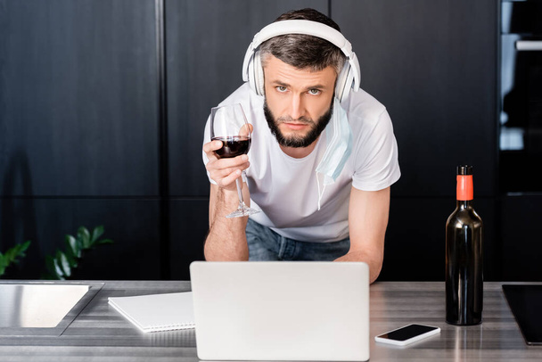 Handsome freelancer in headphones and medical mask looking at camera while holding glass of wine near gadgets on worktop in kitchen  - Photo, Image