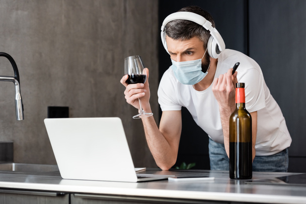 Pensive man in headphones and medical mask holding usb flash drive and glass of wine near laptop in kitchen  - Фото, изображение