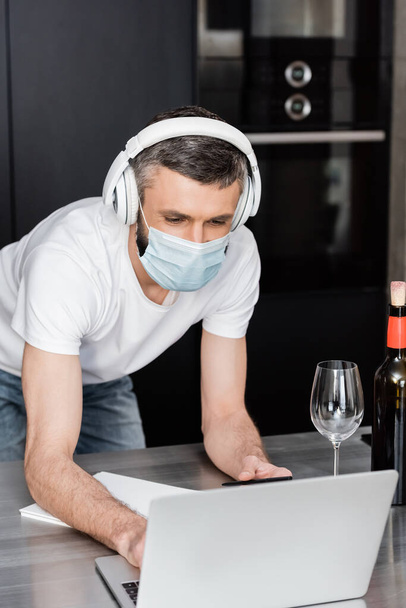 Freelancer in medisch masker en koptelefoon met laptop in de buurt van glas wijn op het werkblad in de keuken  - Foto, afbeelding
