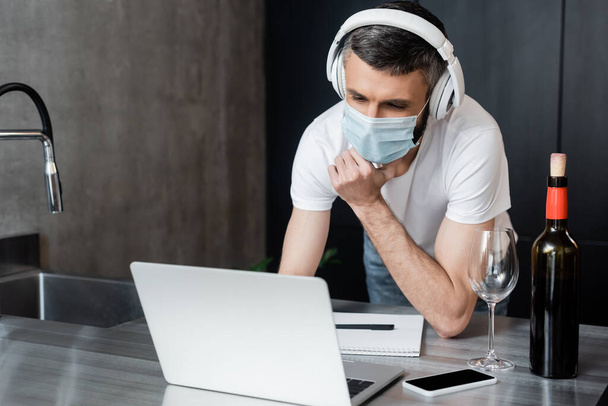Szabadúszó fülhallgató és orvosi maszk nézi laptop közelében bor és okostelefon konyha munkalap  - Fotó, kép