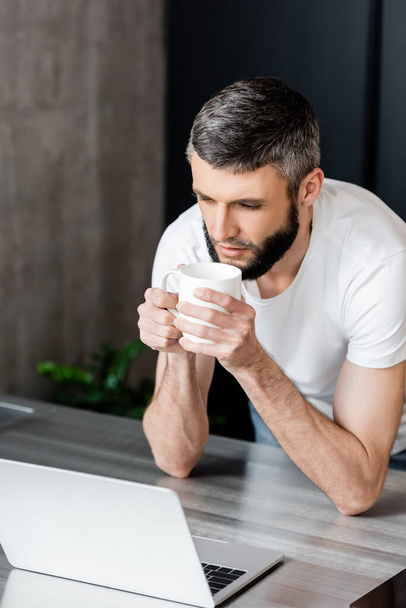 Handsome man holding cup and looking at laptop on kitchen worktop  - Foto, imagen