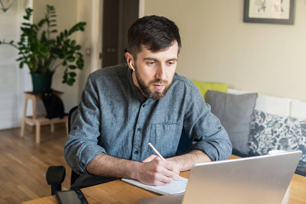 Handsome Eastern European man studying remotely at home during Corona virus quarantine lockdown, writing down during online courses - Photo, Image