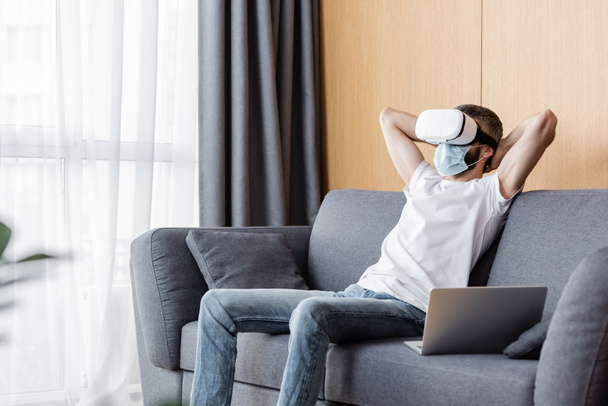 医療用マスクと自宅でソファに座っている仮想現実ヘッドセットの男の選択的な焦点  - 写真・画像