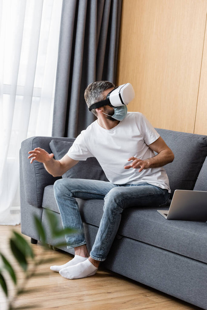 Selective focus of man in medical mask using vr headset on sofa in living room - Foto, afbeelding