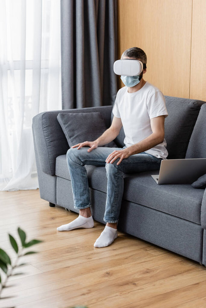 Selective focus of man in medical mask and vr headset sitting near laptop on couch  - Fotó, kép