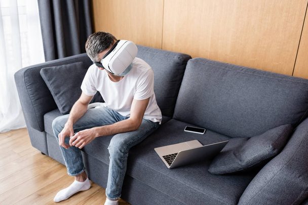 Человек в гарнитуре и медицинской маске сидит рядом со смартфоном и ноутбуком на диване
  - Фото, изображение