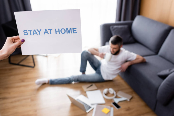 Selective focus of woman holding card with stay at home lettering near man sitting near laptop and stationery on floor at home  - Photo, Image