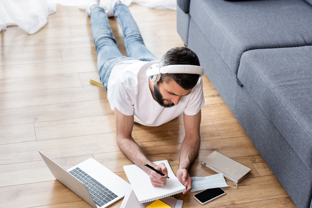 Overhead view van de mens in hoofdtelefoon schrijven op notebook tijdens webinar in de buurt van laptop op de vloer thuis  - Foto, afbeelding