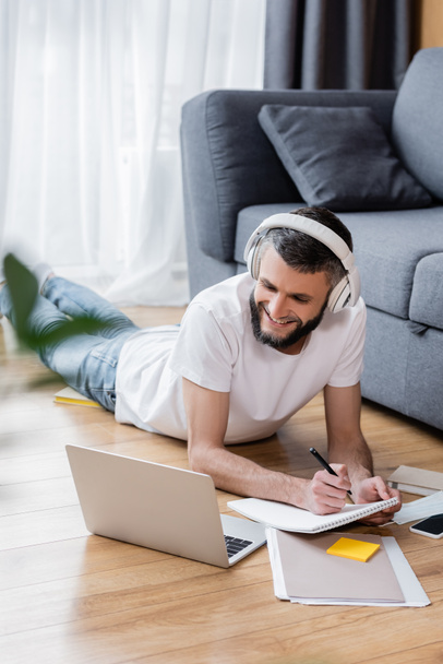 Selective focus of smiling man in headphones using laptop and stationery during online education at home - Fotó, kép