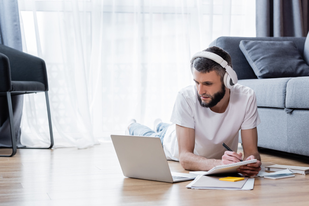 Man in headphones writing on notebook and looking at laptop during webinar in living room - Fotó, kép