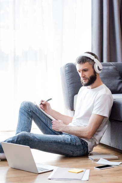 Oldalnézet a férfi fülhallgató írás notebook közelében laptop és írószer az online oktatás során otthon - Fotó, kép