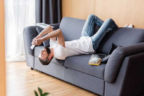 Side view of man using smartphone near books and laptop on sofa at home  - Valokuva, kuva