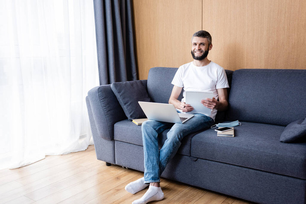 Smiling freelancer holding notebook near laptop, books and medical mask on couch at home  - Photo, Image